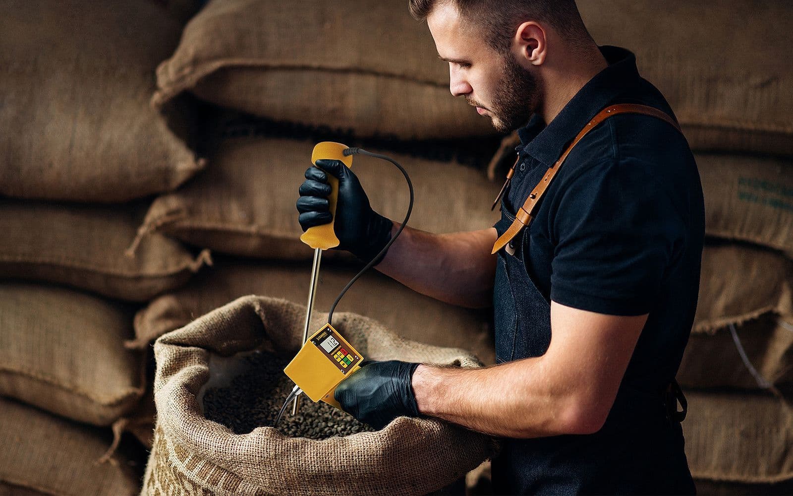 Operario de control de calidad realizando medición de humedad con medidor portátil amarillo sobre saco de yute con granos de café arábica en almacén de recepción